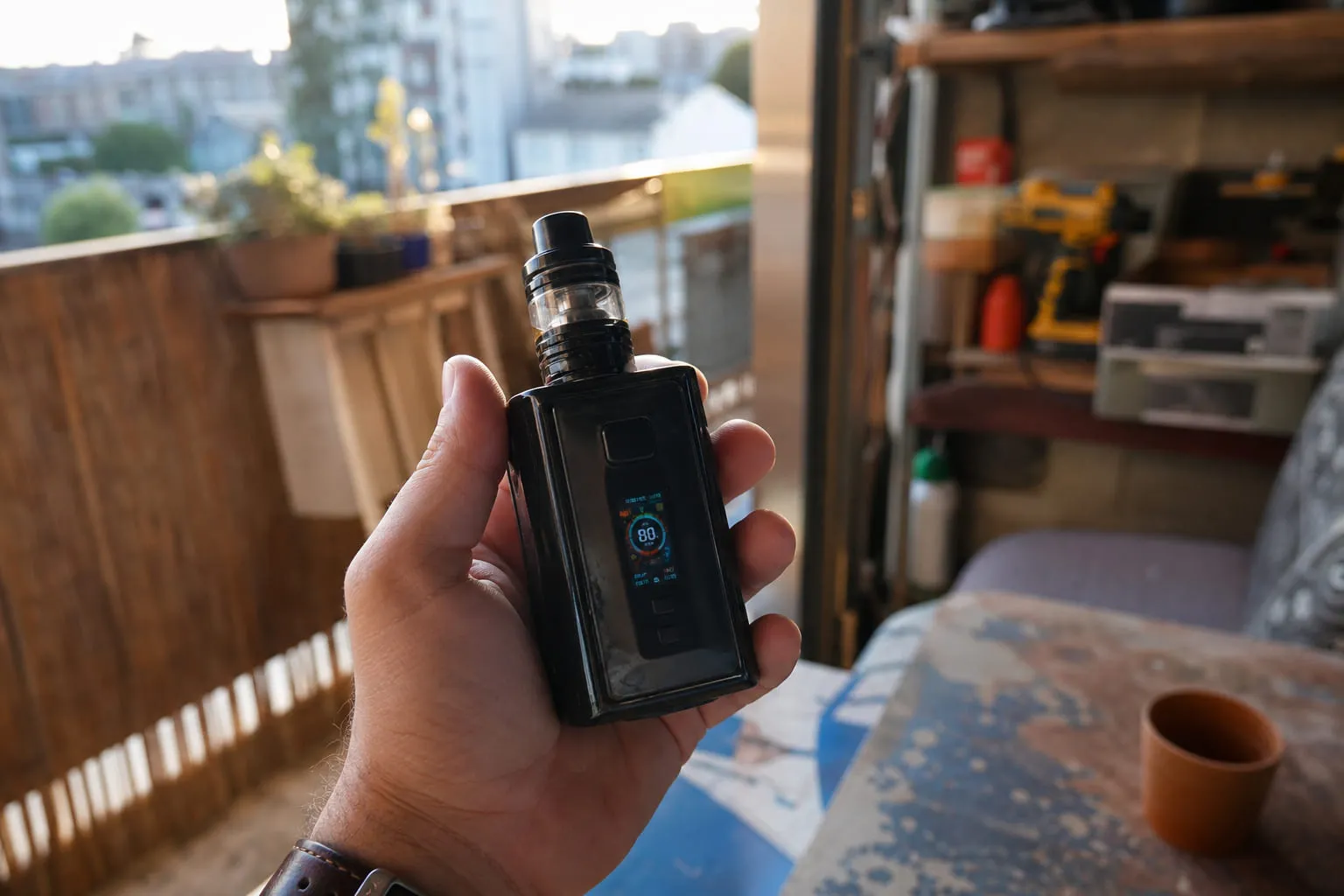 Une box mod avec écran allumé tenue dans une main, atelier en arrière-plan