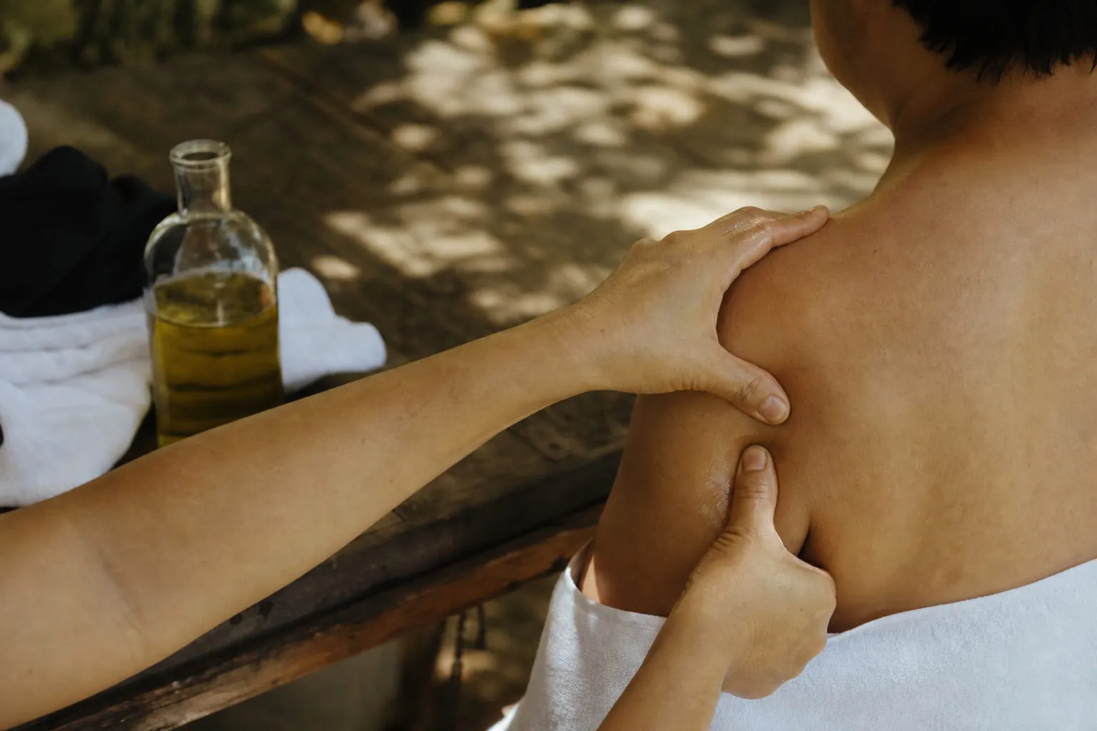 Un flacon d'huile de massage et des mains massant une épaule, serviette blanche