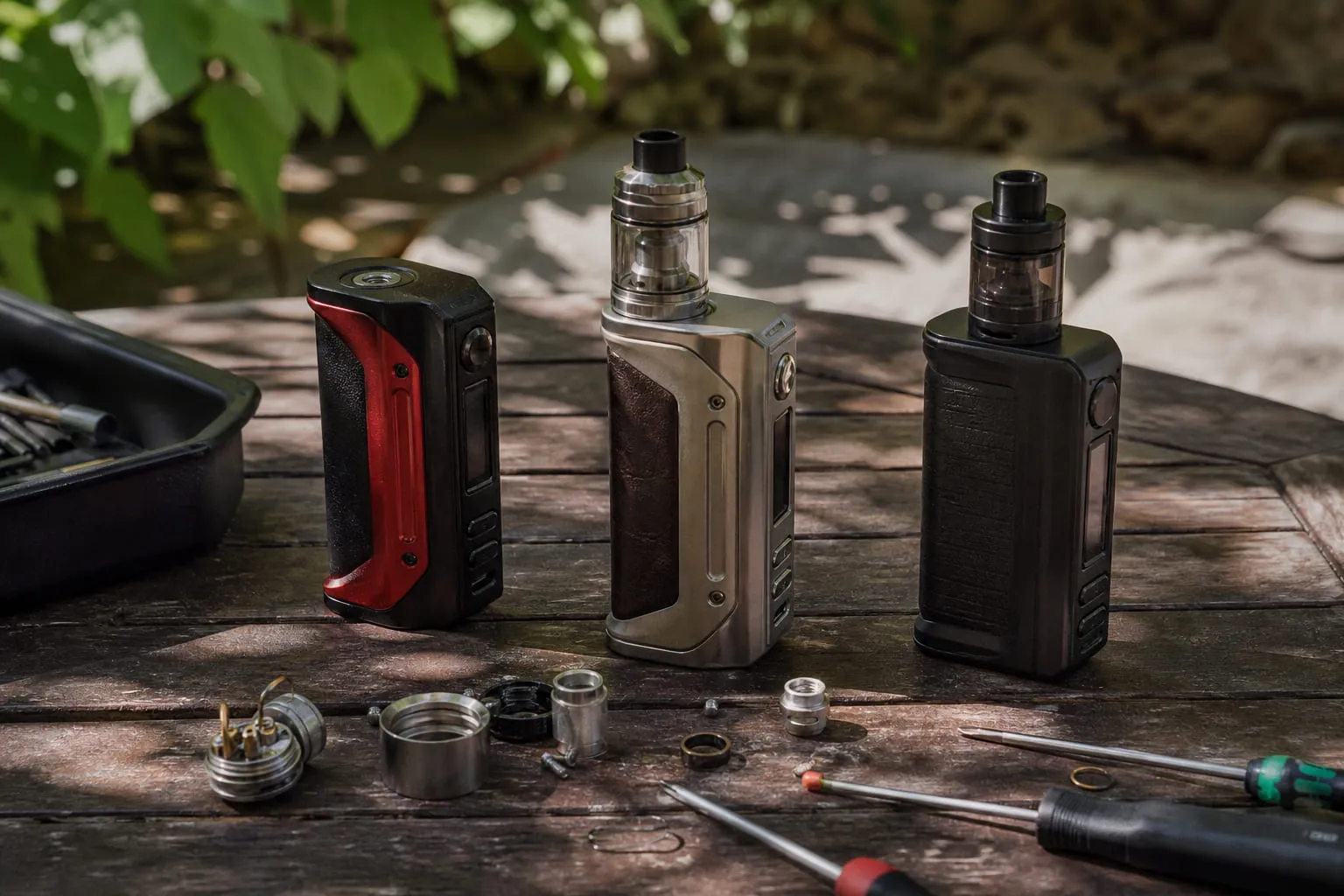 Trois box mods électroniques alignés sur un établi avec des pièces détachées